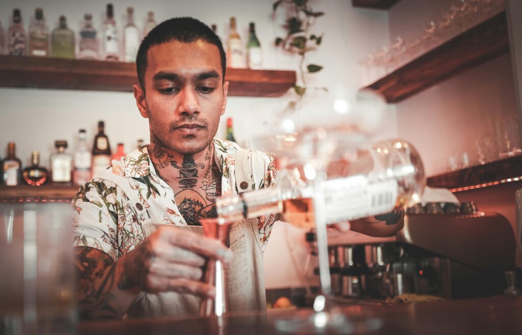 A tattooed bartender pours a drink in a cozy bar setting, showcasing mixology skills.