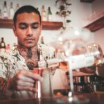 A tattooed bartender pours a drink in a cozy bar setting, showcasing mixology skills.