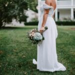 A bride in a white gown stands outdoors holding a bouquet in a lush garden setting.