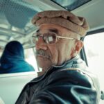 Elderly man with glasses and hat in a vehicle interior, looking back at the camera.