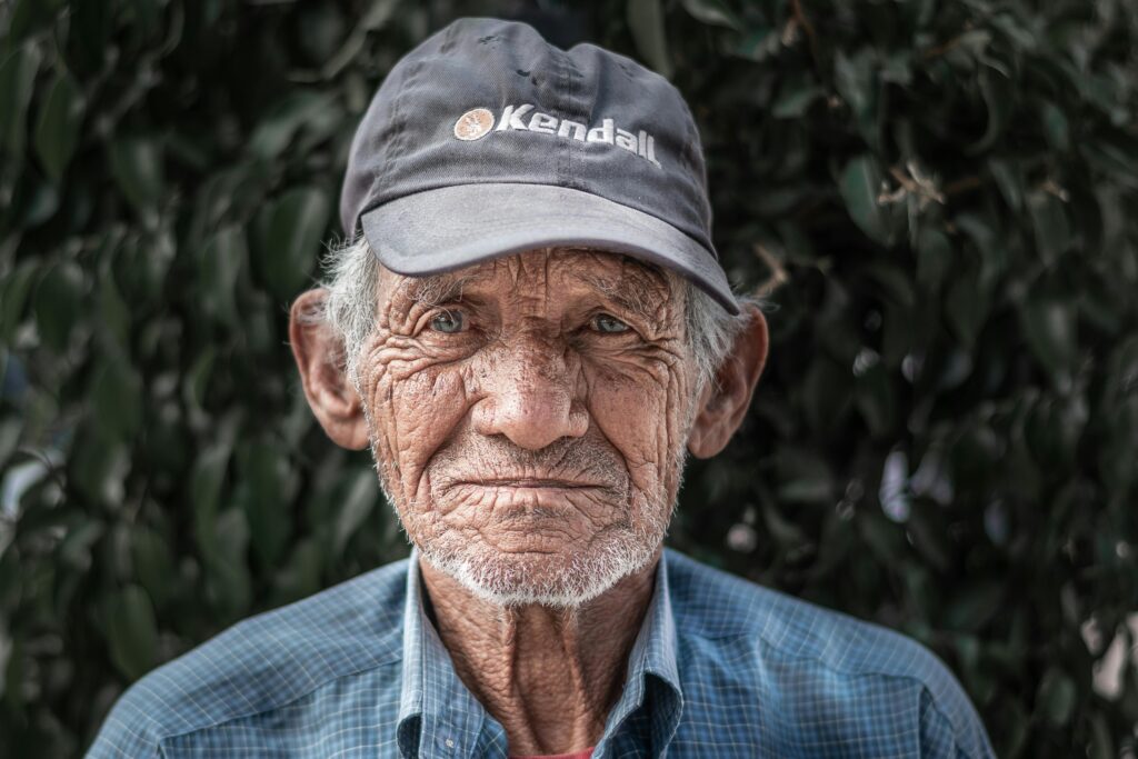 Close-up portrait of an elderly man wearing a cap, outdoors against foliage.