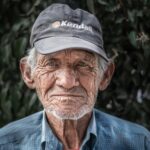 Close-up portrait of an elderly man wearing a cap, outdoors against foliage.