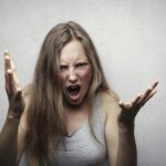 A young woman showing intense anger and frustration, yelling with hands raised against a gray background.