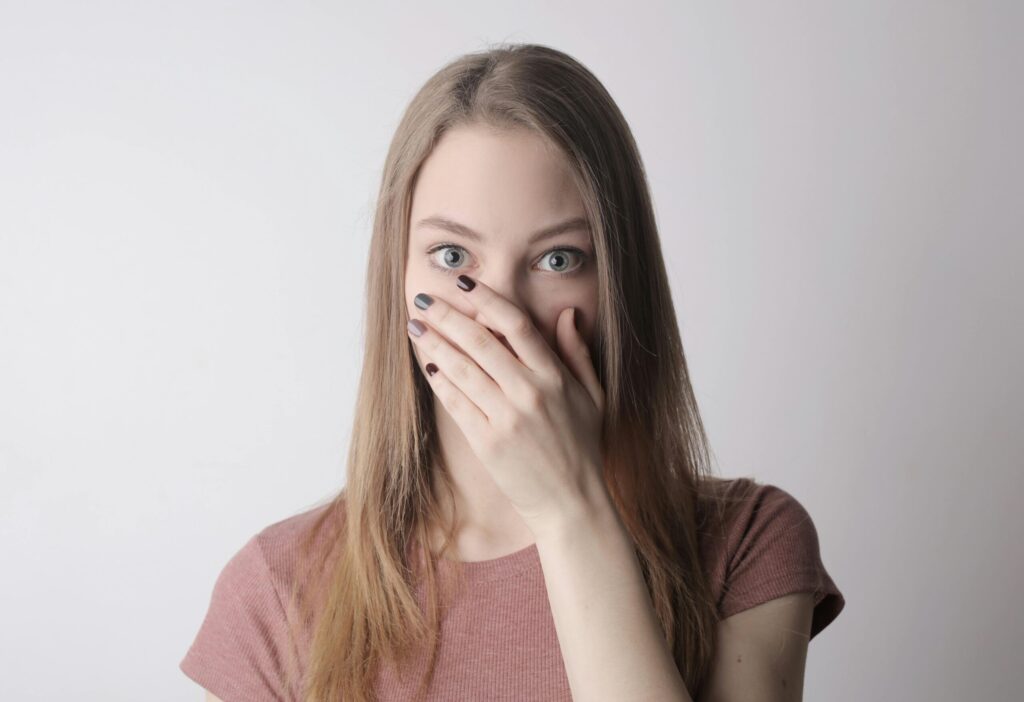 Young woman in pink shirt expressing shock with hand over mouth on simple gray background.