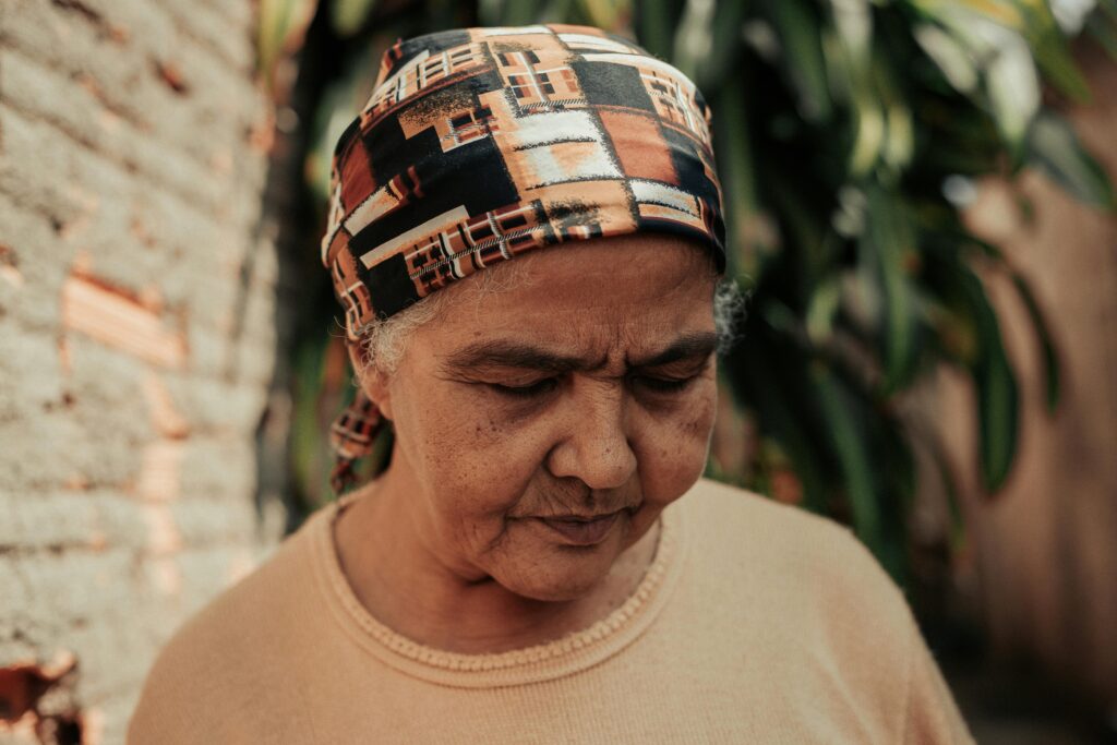 A thoughtful elderly woman in a patterned headscarf stands outdoors, surrounded by greenery.