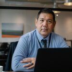 Confident ethnic male entrepreneur using laptop during business work in modern workspace and looking at camera