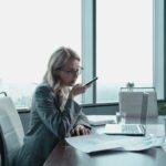 Businesswoman using phone in modern office with laptop and documents.