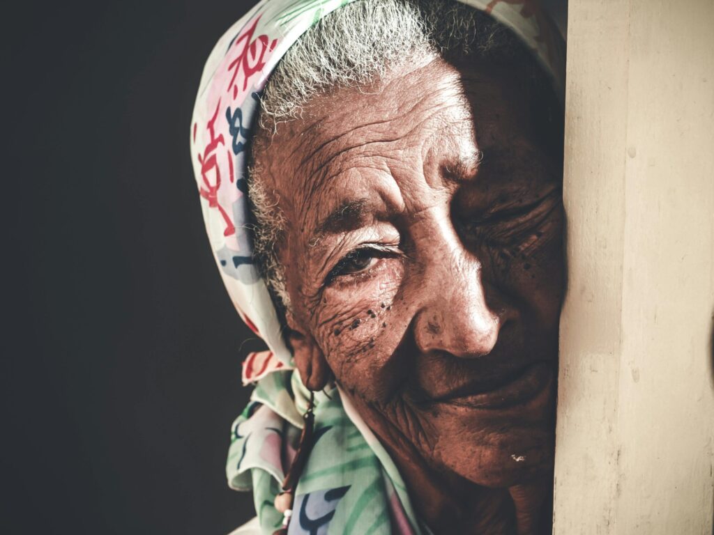 Crop calm senior Asian female with wrinkled face wearing white headscarf standing against black background and looking at camera