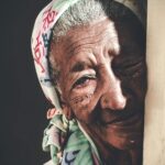 Crop calm senior Asian female with wrinkled face wearing white headscarf standing against black background and looking at camera