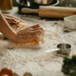 A baker's hands knead dough on a floured kitchen table surrounded by ingredients.