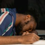 A man with facial hair rests his head on a desk, napping during a late work session.