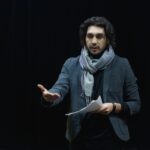 Actor rehearsing on a dark theater stage, holding a script and gesturing dynamically.