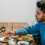 Side view of ethnic male in blue hoodie eating dessert and tasty meal at wooden table