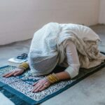 A woman in traditional clothing bowing in prayer on a prayer rug indoors.
