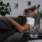 Elderly man lying on a couch with blanket, appearing unwell indoors.