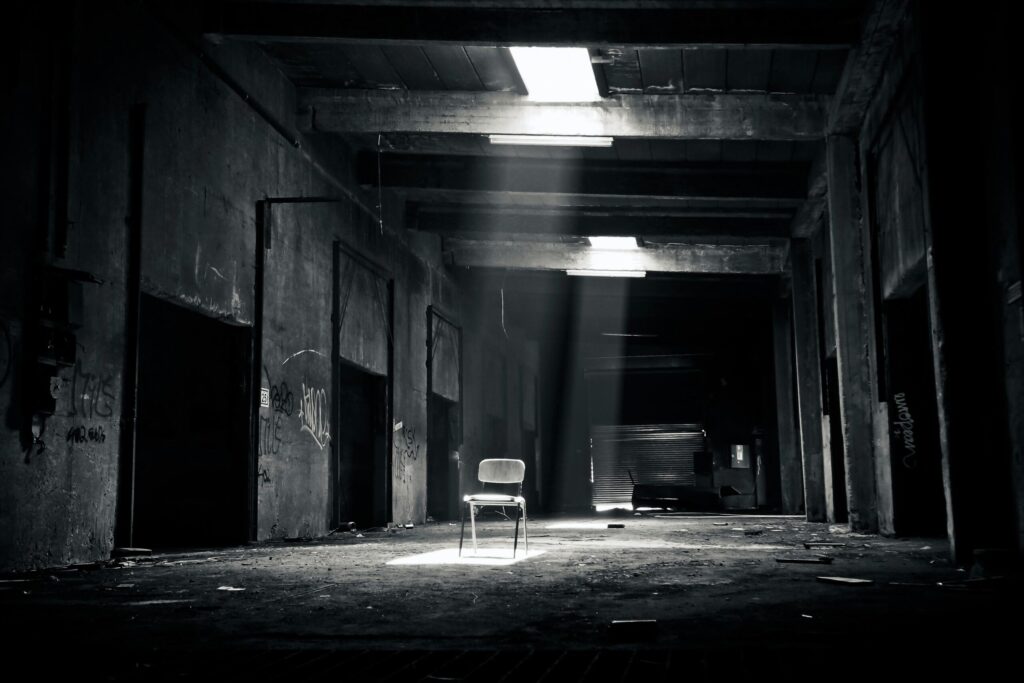 A haunting black and white photo of an abandoned warehouse with a solitary chair illuminated by sunlight through a window.
