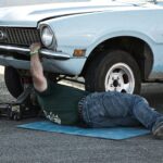 A mechanic working under a classic car on the pavement during the day.