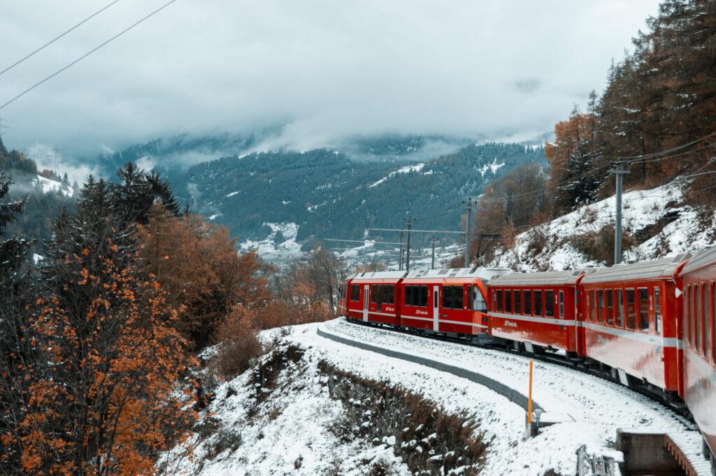 Red train travels through snowy mountains in winter landscape with fog and fall foliage.