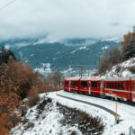 Red train travels through snowy mountains in winter landscape with fog and fall foliage.