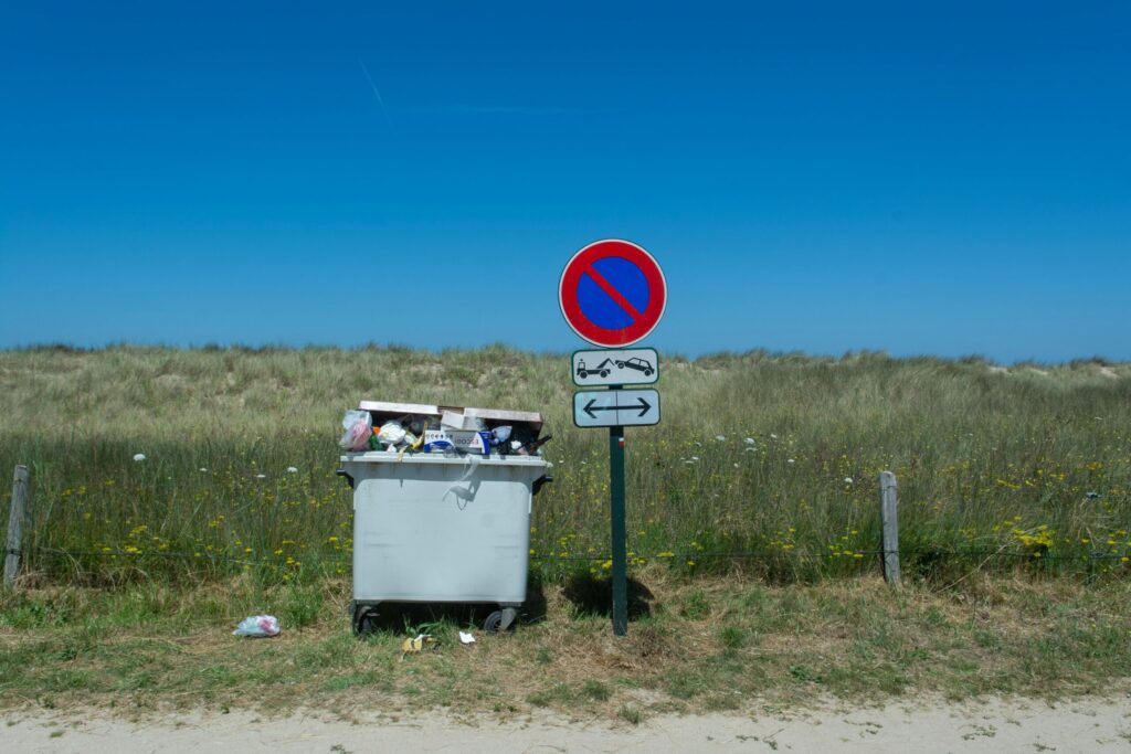 A full garbage bin next to a no parking sign in a countryside setting under clear skies.