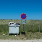 A full garbage bin next to a no parking sign in a countryside setting under clear skies.