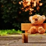 Cute teddy bear sitting on a wooden bench in a sunny, lush garden setting.