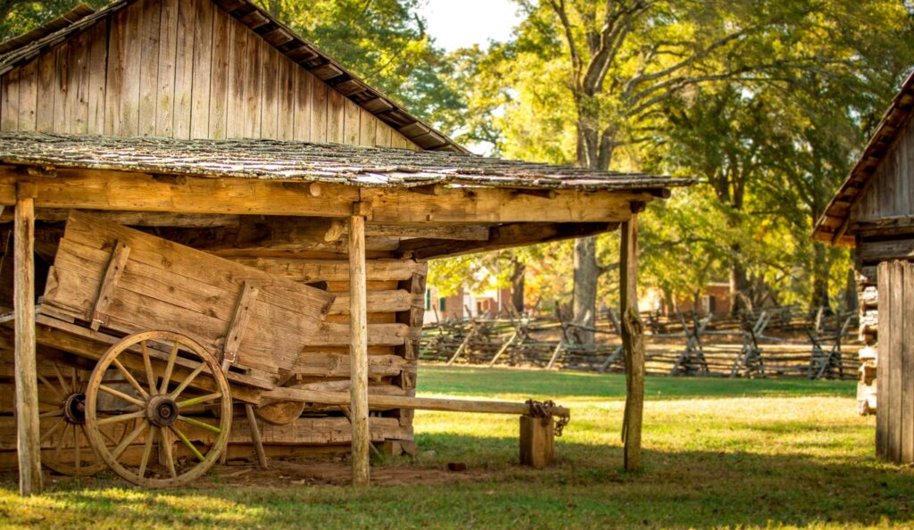A rustic wooden barn with an old wooden wagon in a sunny outdoor farm setting.