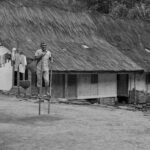 Black and white photo of a man on stilts in front of a thatched-roof house.