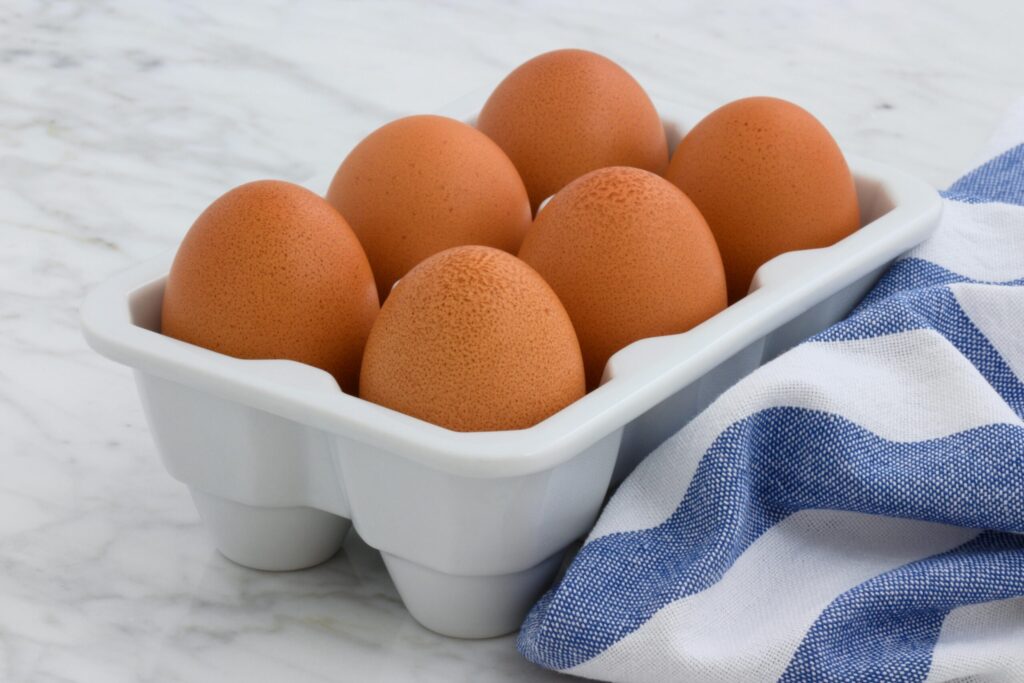 Six fresh brown eggs in a white tray on a marble surface, with a blue towel.