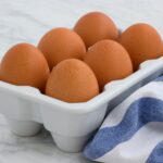 Six fresh brown eggs in a white tray on a marble surface, with a blue towel.