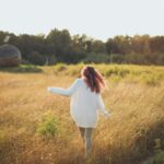 Woman in a white sweater walking through a sunlit grassy field, embracing nature and tranquility.