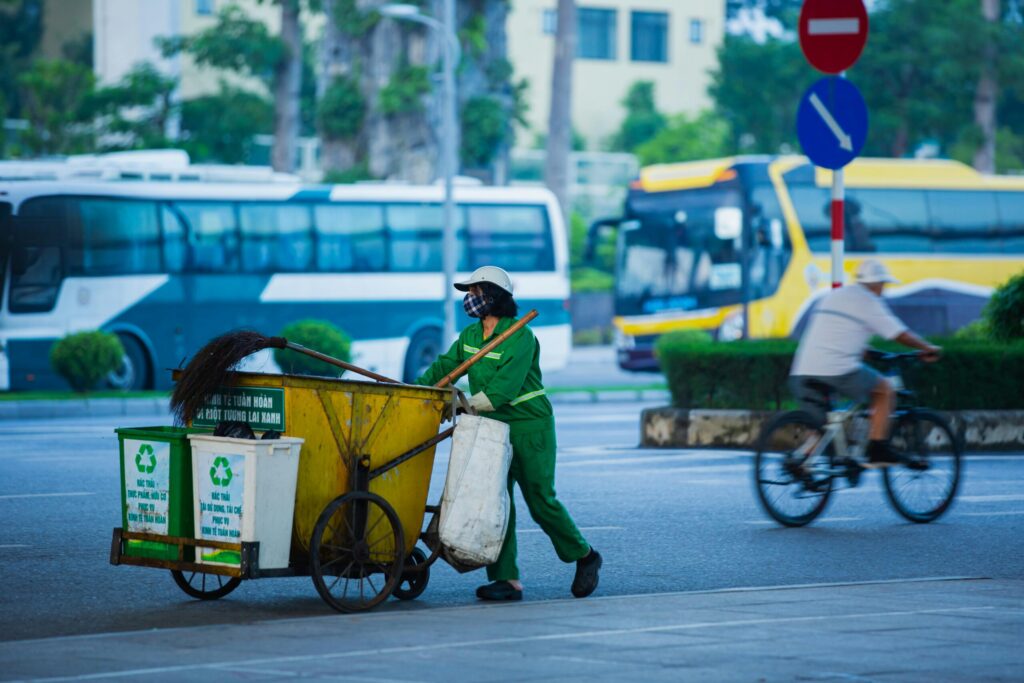 Street cleaner pushing cart with recycling bins in a busy urban setting.