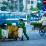 Street cleaner pushing cart with recycling bins in a busy urban setting.