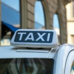 Close-up view of a taxi sign atop a vehicle on an urban city street with blurred architectural background.