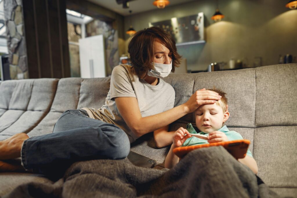 A concerned mother wearing a mask checks her child's temperature on the sofa.