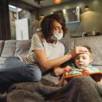 A concerned mother wearing a mask checks her child's temperature on the sofa.