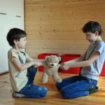 Side view full length irritated fighting brothers sitting on floor and pulling teddy bear to sides