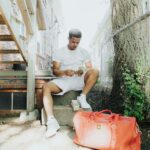 A young man counting money while sitting outside near a staircase with a bold orange duffel bag.