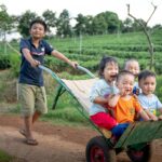 Five children having fun on a wheelbarrow ride in a rural field, enjoying the outdoors.