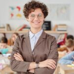 Smiling teacher with glasses standing with arms crossed in a classroom, students learning.