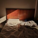 A sunlit, unmade bed with a wooden headboard creates a cozy and warm ambience.