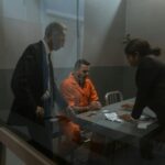 A tense interrogation in progress with a handcuffed inmate and two officers in a prison room.