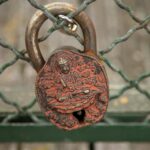 Close-up of an ornately designed rusty padlock hanging on a metal fence, depicting intricate engraved patterns.