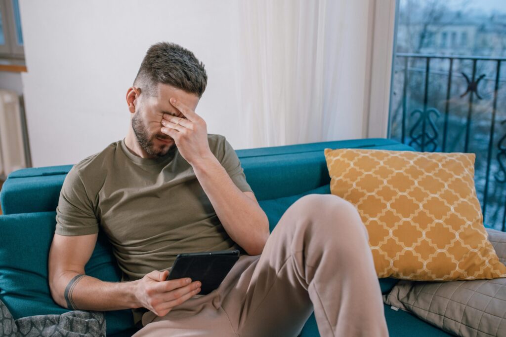 A man sits on a sofa holding a tablet, visibly stressed and covering his face with one hand.