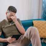 A man sits on a sofa holding a tablet, visibly stressed and covering his face with one hand.