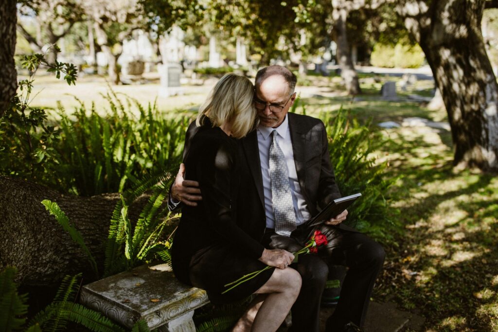 A couple dressed in black sitting on a bench in a cemetery, holding a rose and a picture frame.