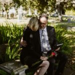 A couple dressed in black sitting on a bench in a cemetery, holding a rose and a picture frame.