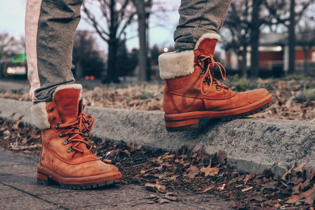 Close-up of orange leather boots with fur lining on a street curb amidst dry leaves, conveying autumn fashion vibes.