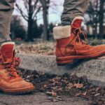 Close-up of orange leather boots with fur lining on a street curb amidst dry leaves, conveying autumn fashion vibes.