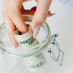A close-up of a hand placing rolled dollars into a glass jar, symbolizing savings.